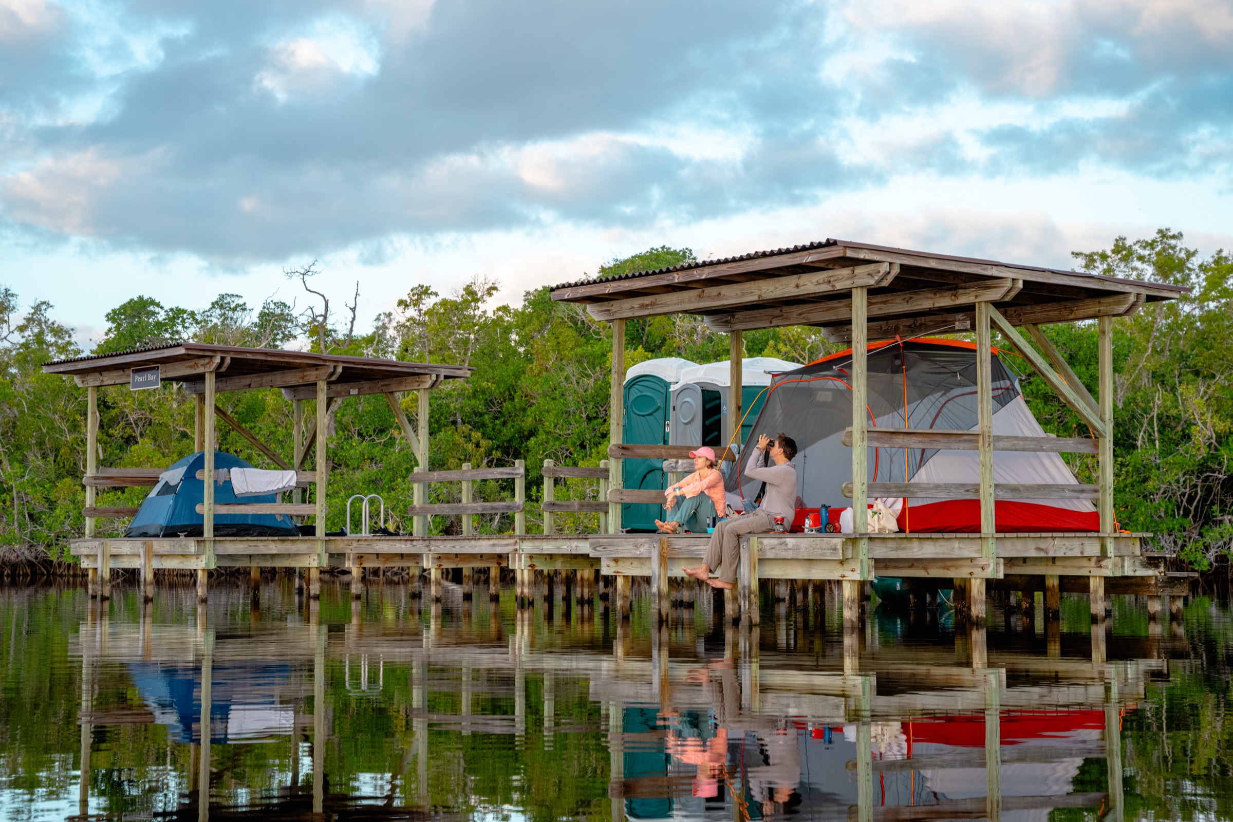 Two tents set up on an elevated wooden chickee platform above the water in the Florida Everglades, surrounded by mangroves, with visitors visible on the structure