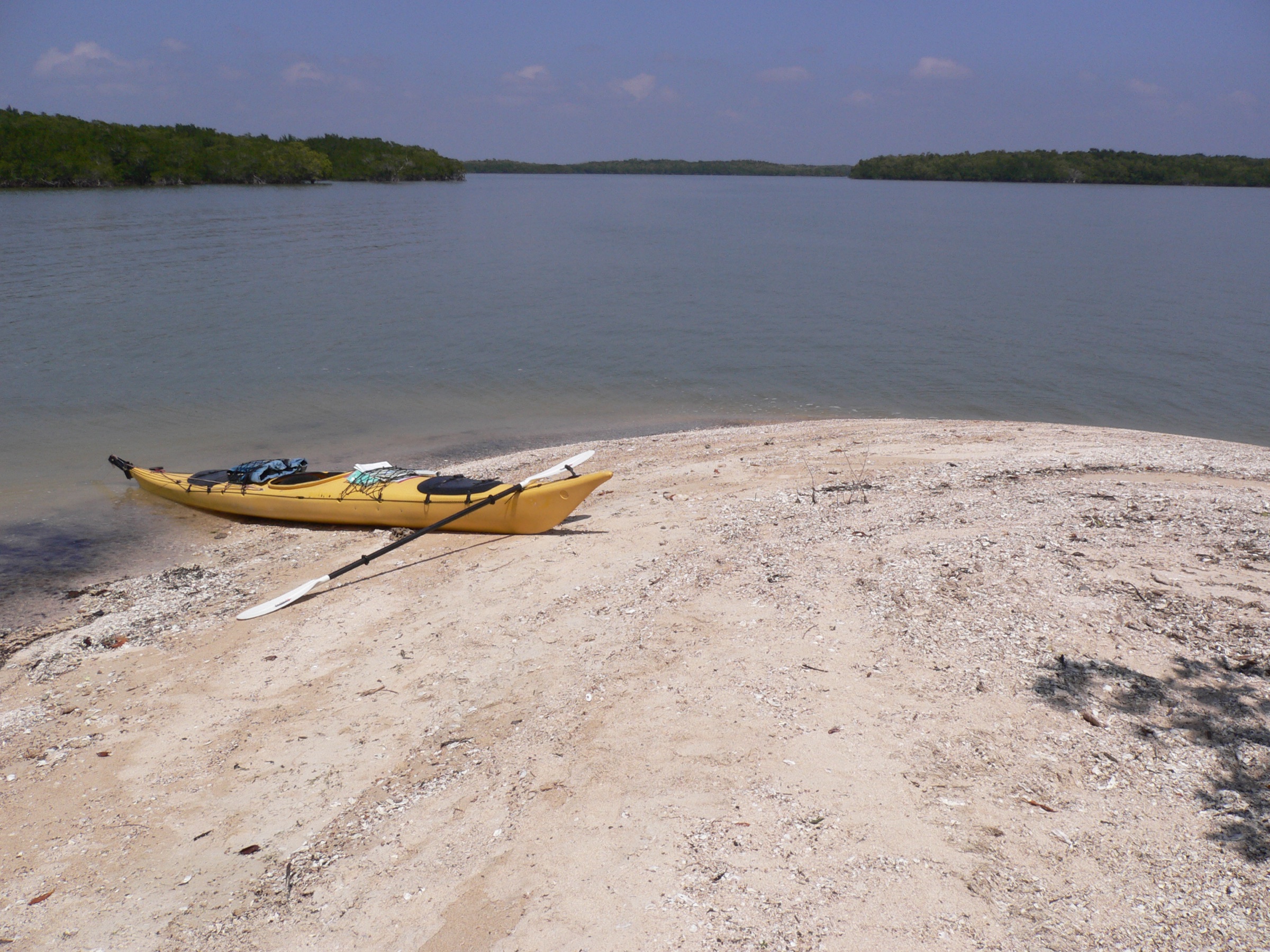 A yellow sea kayak loaded with gear rests on a shell-sand beach in Florida's Ten Thousand Islands, with calm water and mangrove islands stretching into the distance